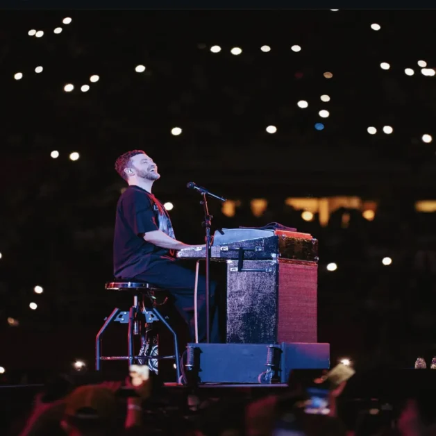 Justin Timberlake plays keyboard on stage under spotlight with crowd lights in the background