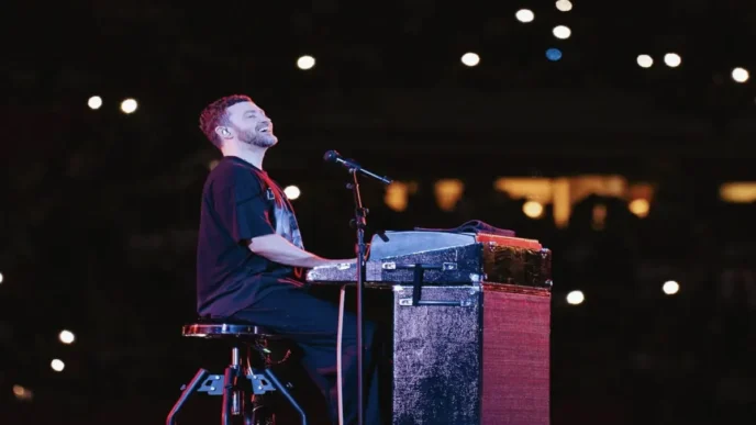 Justin Timberlake plays keyboard on stage under spotlight with crowd lights in the background