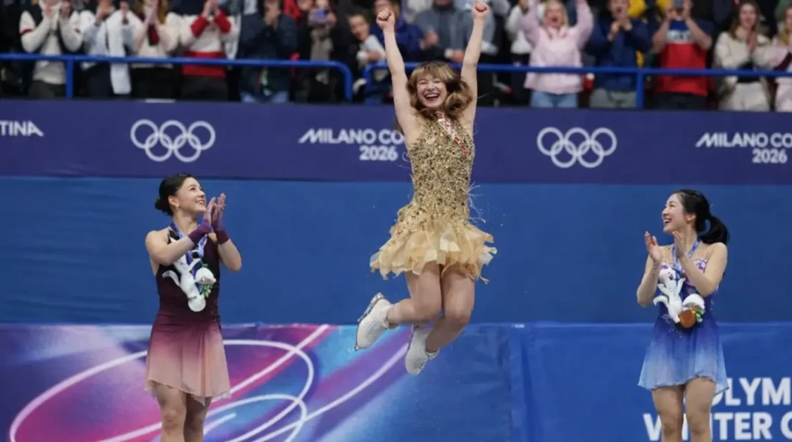 Alysa Liu celebrates on the Olympic podium after figure skating, jumping with arms raised as rivals applaud.