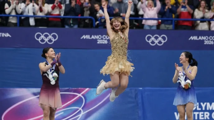 Alysa Liu celebrates on the Olympic podium after figure skating, jumping with arms raised as rivals applaud.