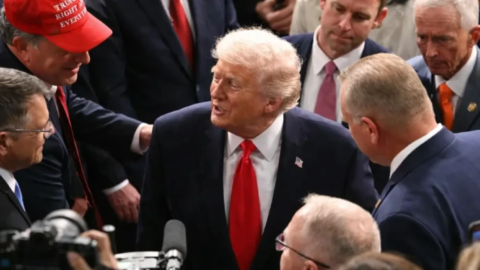 Donald Trump in a navy suit and red tie speaks with people while cameras and microphones surround him at a crowded event