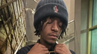 Luci4 wearing a dark beanie and adjusting a gold chain necklace in an indoor setting