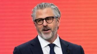 Casey Wasserman in navy suit and glasses speaking at podium against a red conference backdrop