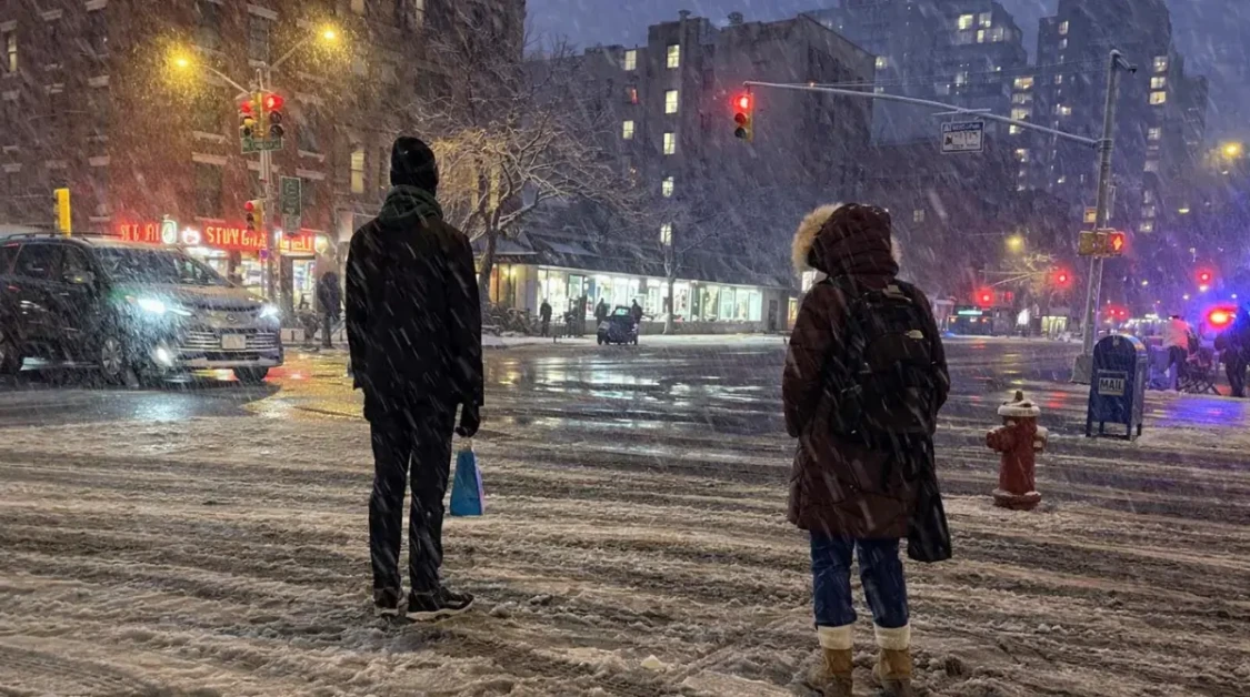 new york city ban scene at a snowy night intersection with pedestrians waiting to cross and traffic lights glowing