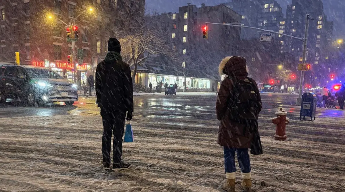 new york city ban scene at a snowy night intersection with pedestrians waiting to cross and traffic lights glowing