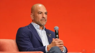 Peter Attia seated on a stage chair holding a microphone against an orange background