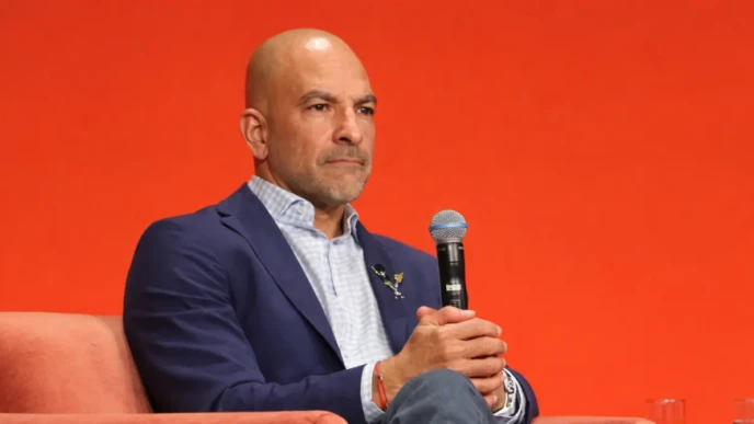 Peter Attia seated on a stage chair holding a microphone against an orange background