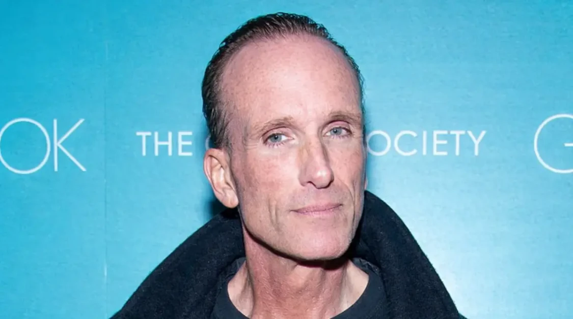 Peter Greene pictured at a public event in a black coat against a blue backdrop.