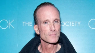 Peter Greene pictured at a public event in a black coat against a blue backdrop.