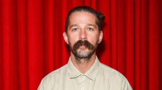Portrait of Shia LaBeouf standing in front of a red curtain wearing a beige shirt