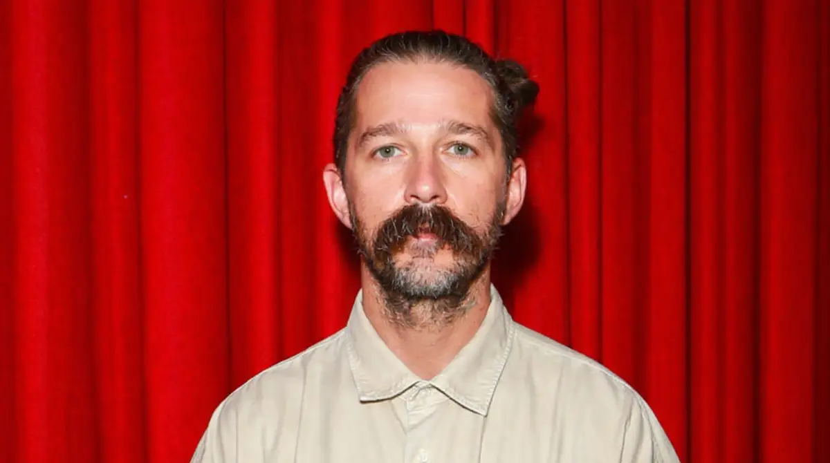 Portrait of Shia LaBeouf standing in front of a red curtain wearing a beige shirt