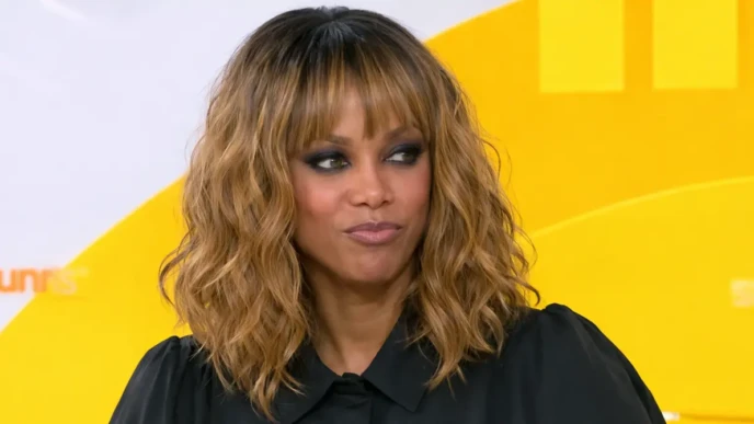 Tyra Banks portrait with wavy blonde-brown hair and black outfit on yellow studio background