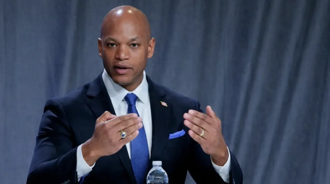 Wes Moore speaking at a panel discussion in a navy suit with hand gestures and a gray backdrop