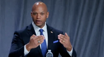 Wes Moore speaking at a panel discussion in a navy suit with hand gestures and a gray backdrop