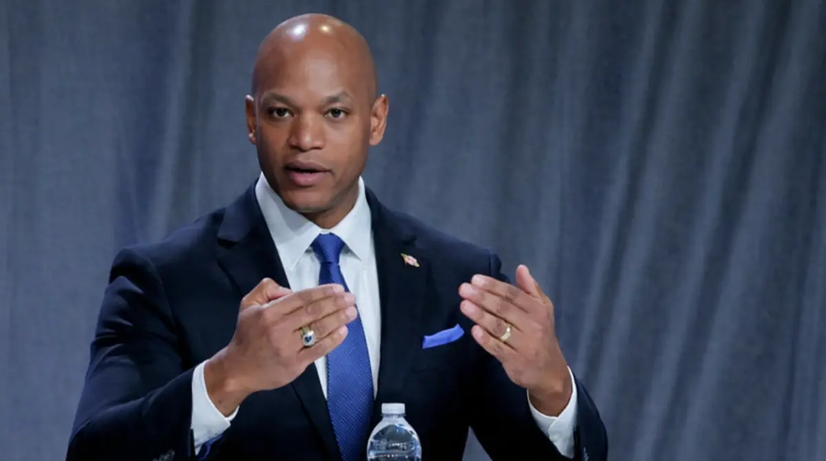 Wes Moore speaking at a panel discussion in a navy suit with hand gestures and a gray backdrop