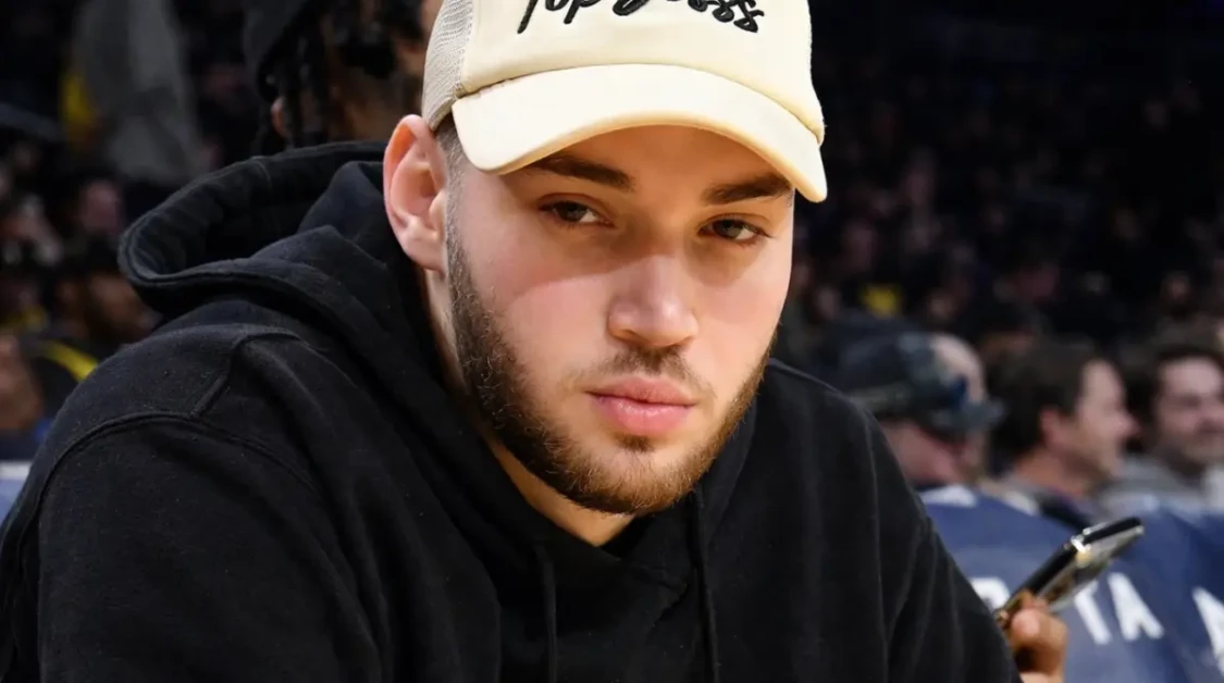 Adin Ross sitting courtside at an arena event wearing a beige cap and black hoodie, looking toward the camera.