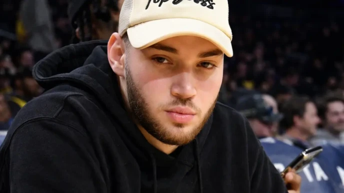 Adin Ross sitting courtside at an arena event wearing a beige cap and black hoodie, looking toward the camera.